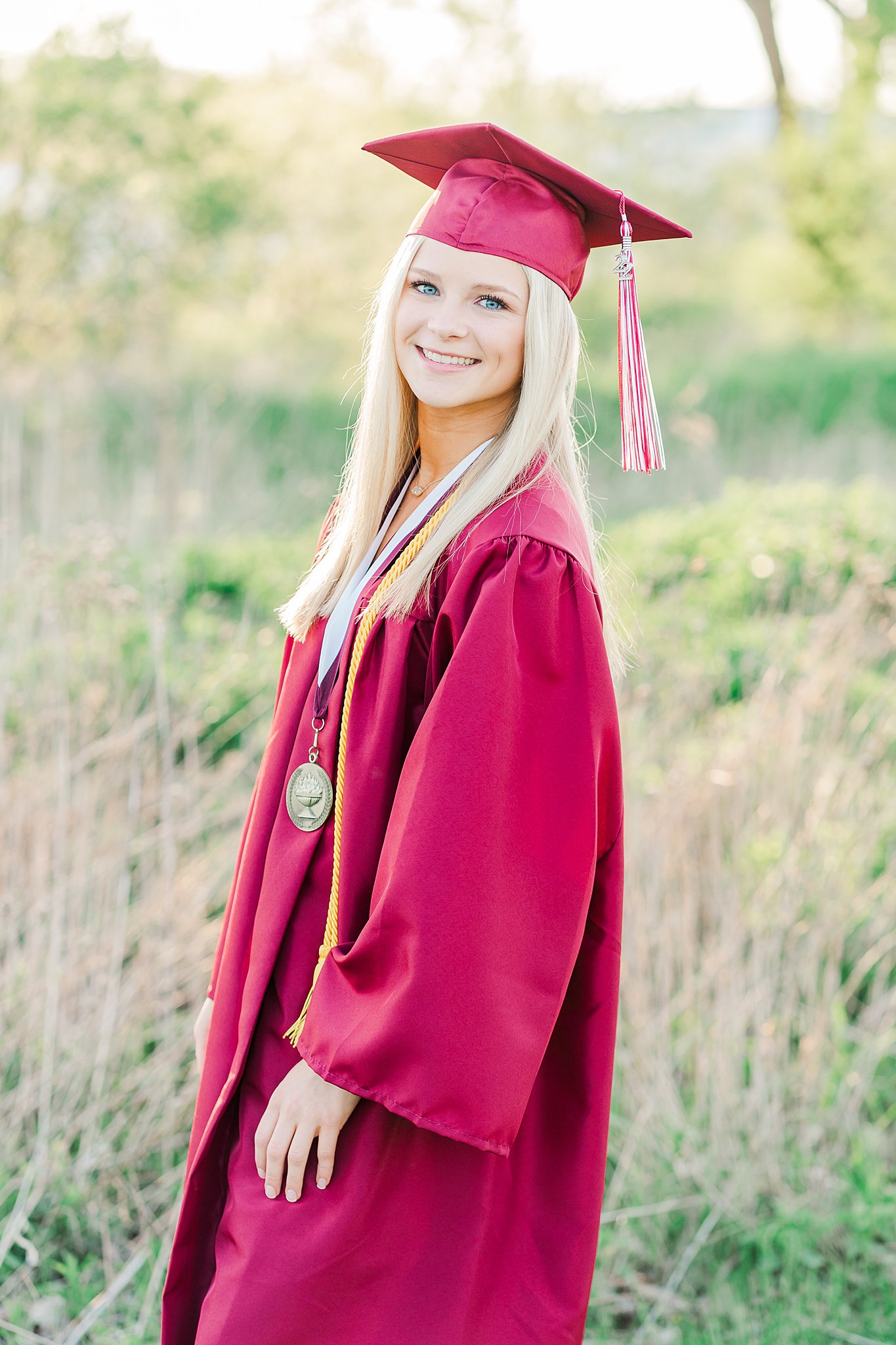 Girl wearing graduation robe and cap stands in a field with tall grass