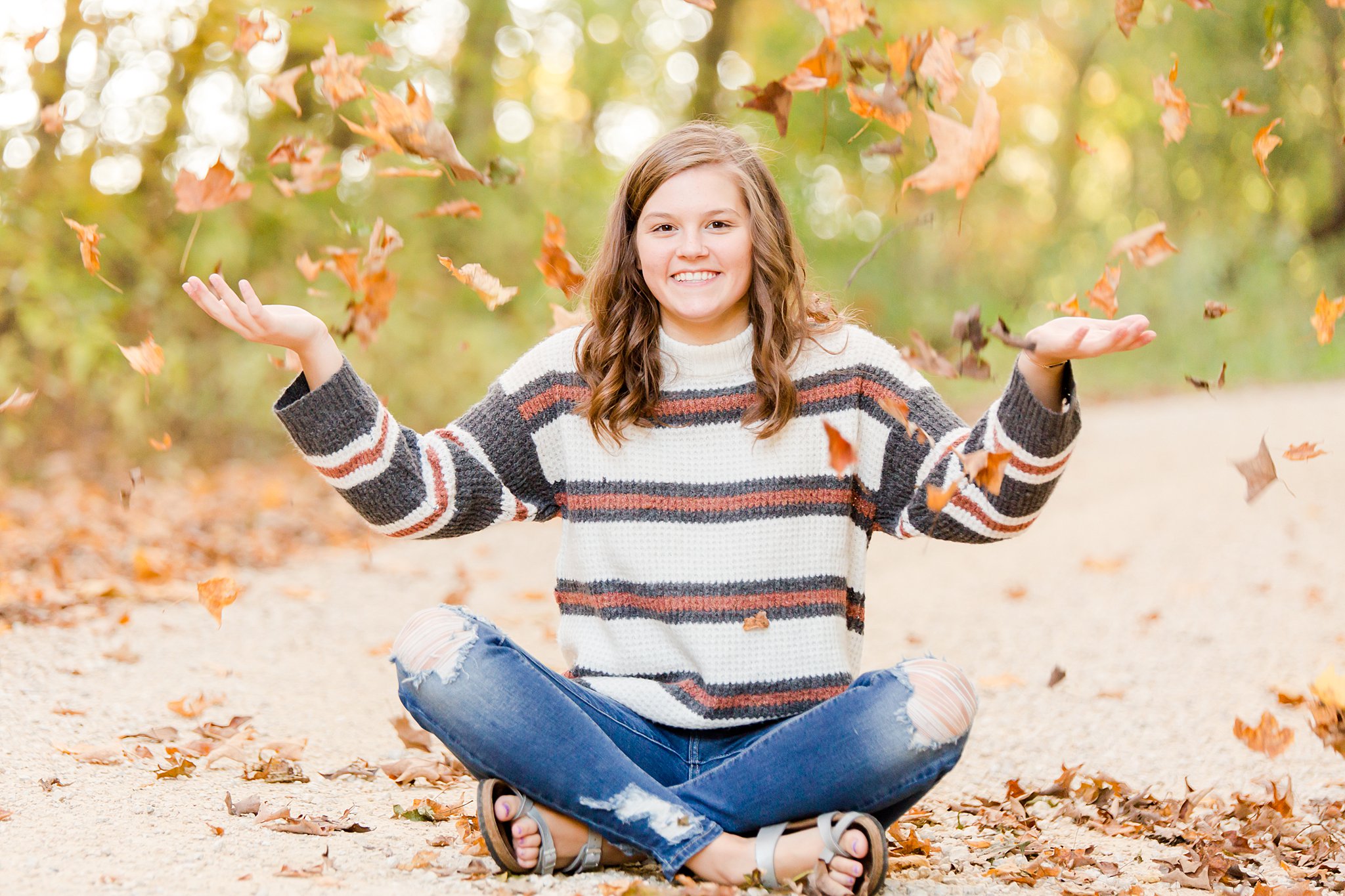 girl sits in jeans and a stripe knit sweater throwing fallen leaves all around her mayo senior high school