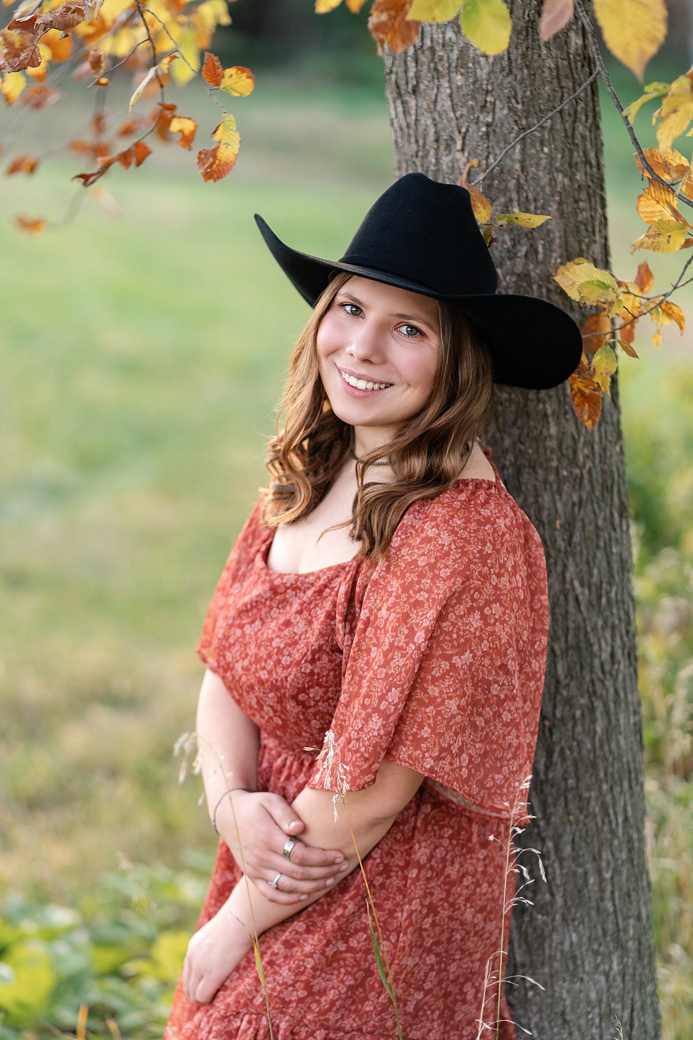 Teenage girl leans against a tree in a field wearing a red pattern dress and black hat stewartville high school